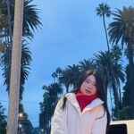 A femme presenting person with long black hair is standing and facing the camera wearing a white jacket and surrounded by palm trees.