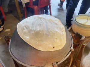 Thin crepe batter cooking on a dome-shaped griddle, with extra batter nearby in a busy setting.