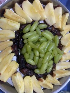 A circular arrangement of sliced apples, dates, and green grapes in a metal tray. The setup appears fresh and inviting.