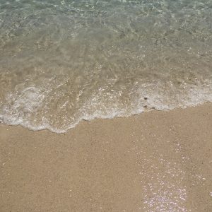 Clear ocean water and white foam washing onto a golden sandy beach. Top-down view of a sunny shoreline with light reflections on the wet sand.