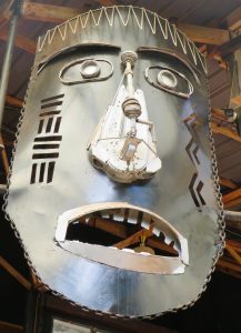 Dark metal face sculpture with mechanical eyes, a rod nose, geometric cutouts, and a jagged mouth, framed by a heavy chain.
