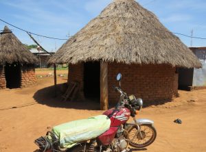 Red motorcycle with a green sack parked on red soil, with two thatched brick huts in the background under a blue sky.