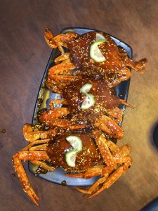 Three crabs covered in spicy red sauce with sesame seeds, garnished with lime slices on a black plate. The tone is appetizing and vibrant.