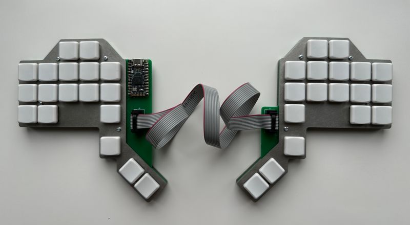 The FG-11 split keyboard. Each side has 23 keys. The keys for the fingers are layed out in five columns or three rows on each side. The second and third column from the inside are one key higher further from the typist than the other columns, to suit where my middle and ring fingers sit. There are three thumb keys on each side, arranged along a diagonal. The PCB of each side can be seen peaking out on the inside edge, and the controller can be seen on the left hand. A ribbon cable connects the two sides from PCB to PCB. The plates are stainless steel and key caps white.