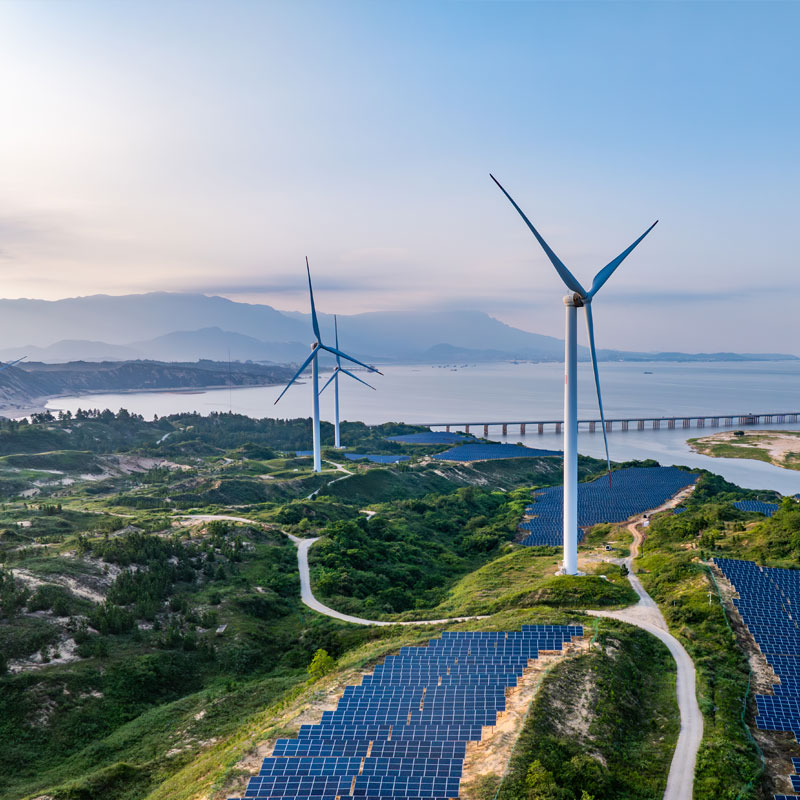 Power-gen-Box-Image Large white wind turbines and rows of solar panels situated on green hills overlooking a bay.